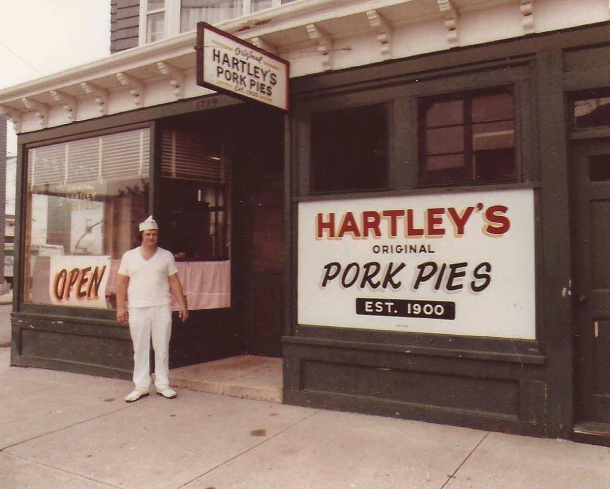Vintage photo of the Hartley's Original Pork Pies storefront, with a baker in white standing by the open door beneath the 'Est. 1900' sign